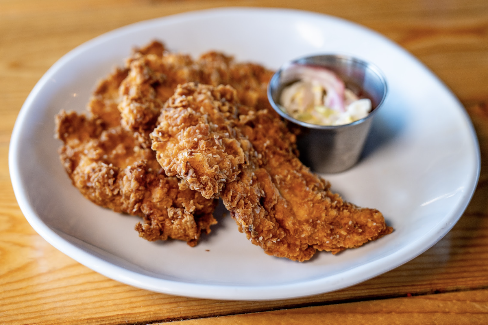 A white plate with crispy fried chicken tenders and a small metal cup of coleslaw sits on a wooden table.