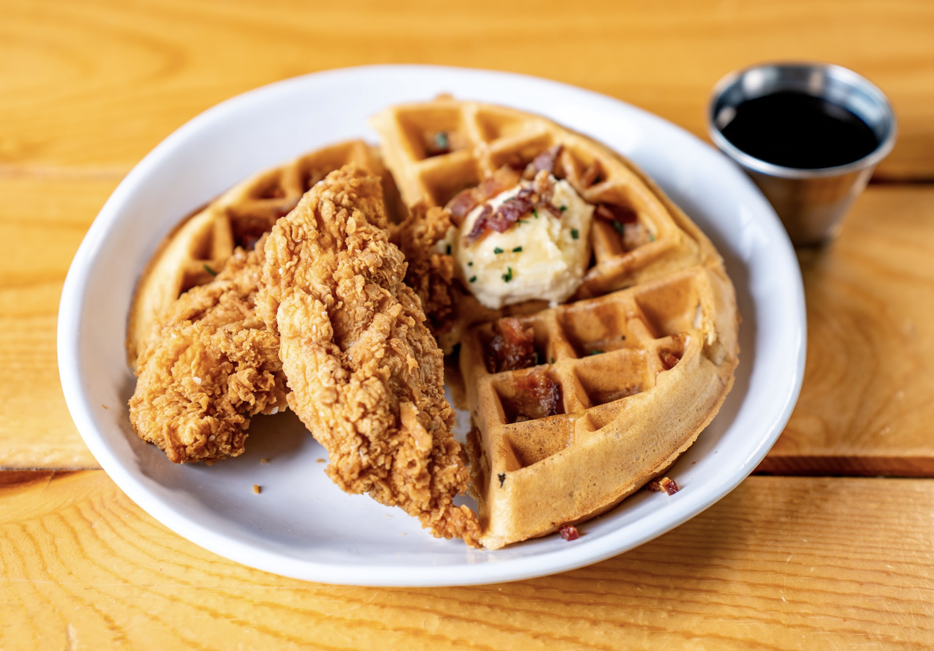 A plate with two pieces of fried chicken on top of a waffle, garnished with butter and chives. A small cup of syrup sits on the side, all placed on a wooden table.