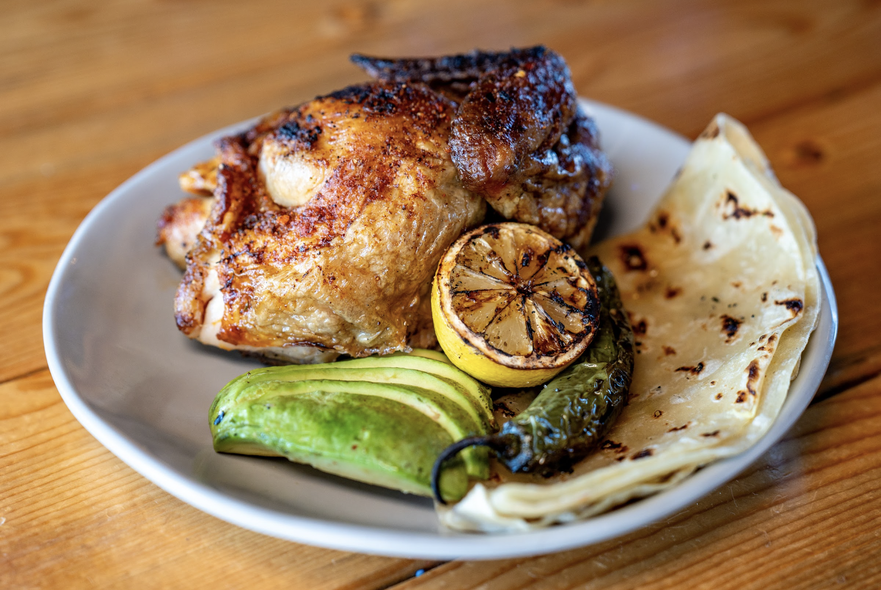 A plate with roasted chicken, grilled lemon, a charred green chili, sliced avocado, and two pieces of grilled flatbread on a wooden table.