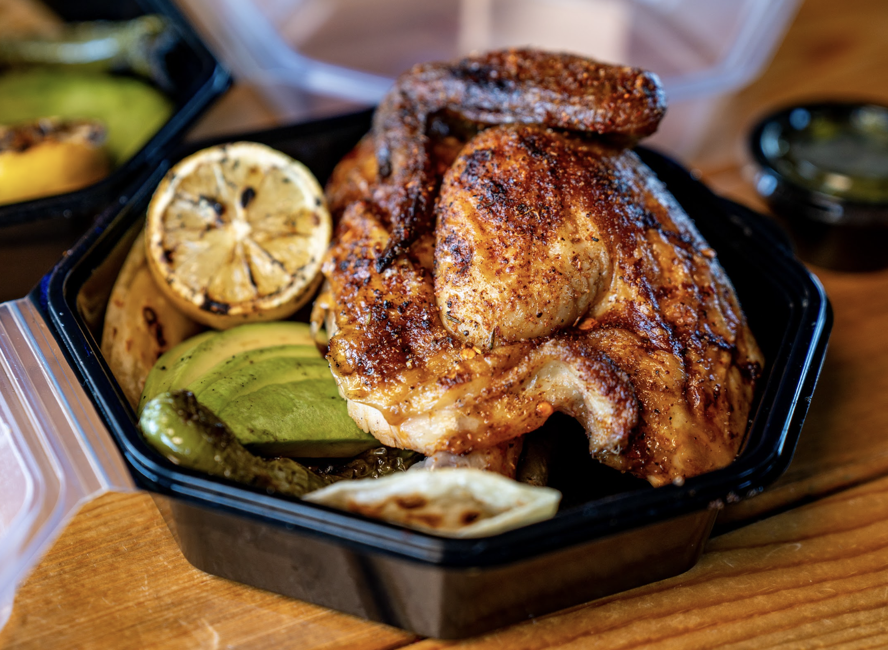 A black takeout container filled with grilled, seasoned chicken, sliced avocado, grilled lemon, jalapeño, and onions on a wooden table.