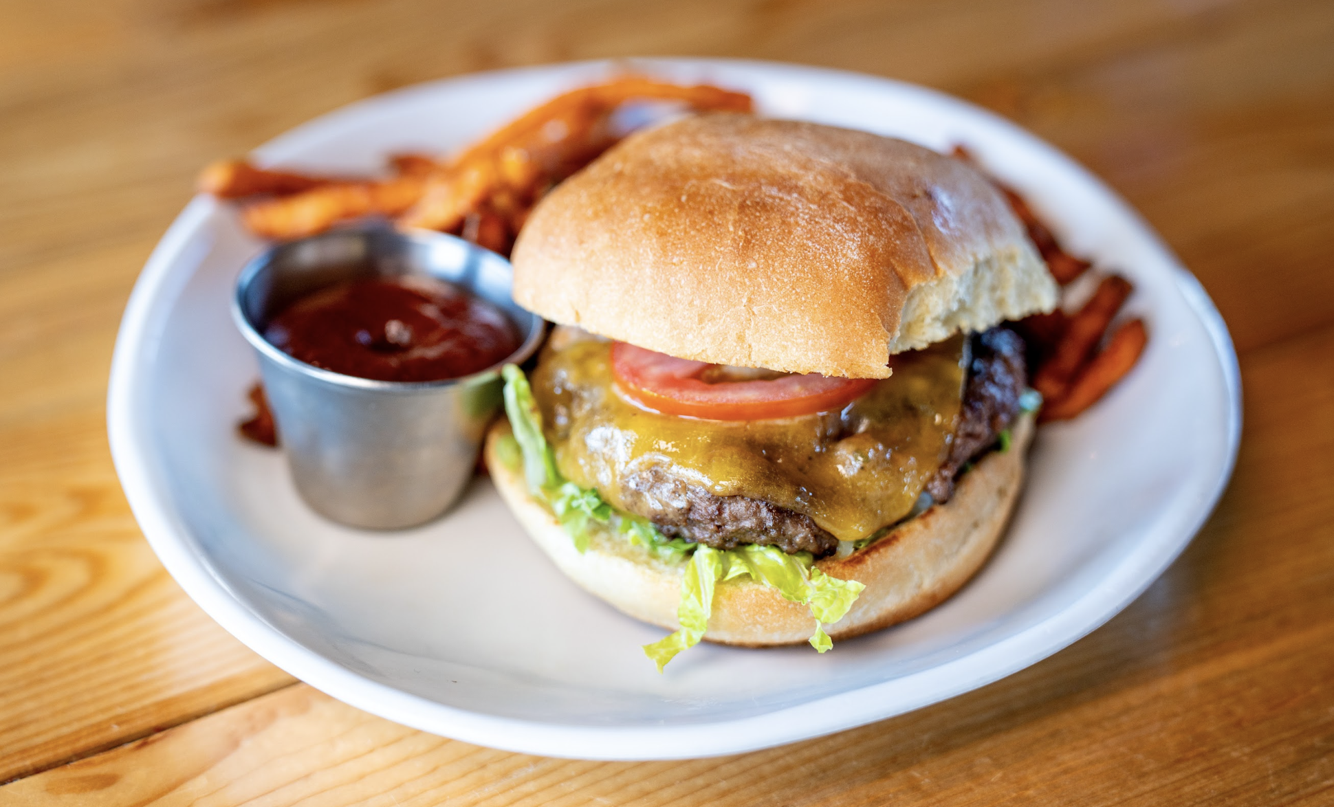 A cheeseburger with lettuce and tomato on a bun, served with sweet potato fries and a small cup of ketchup on a white plate. The plate is on a wooden table.