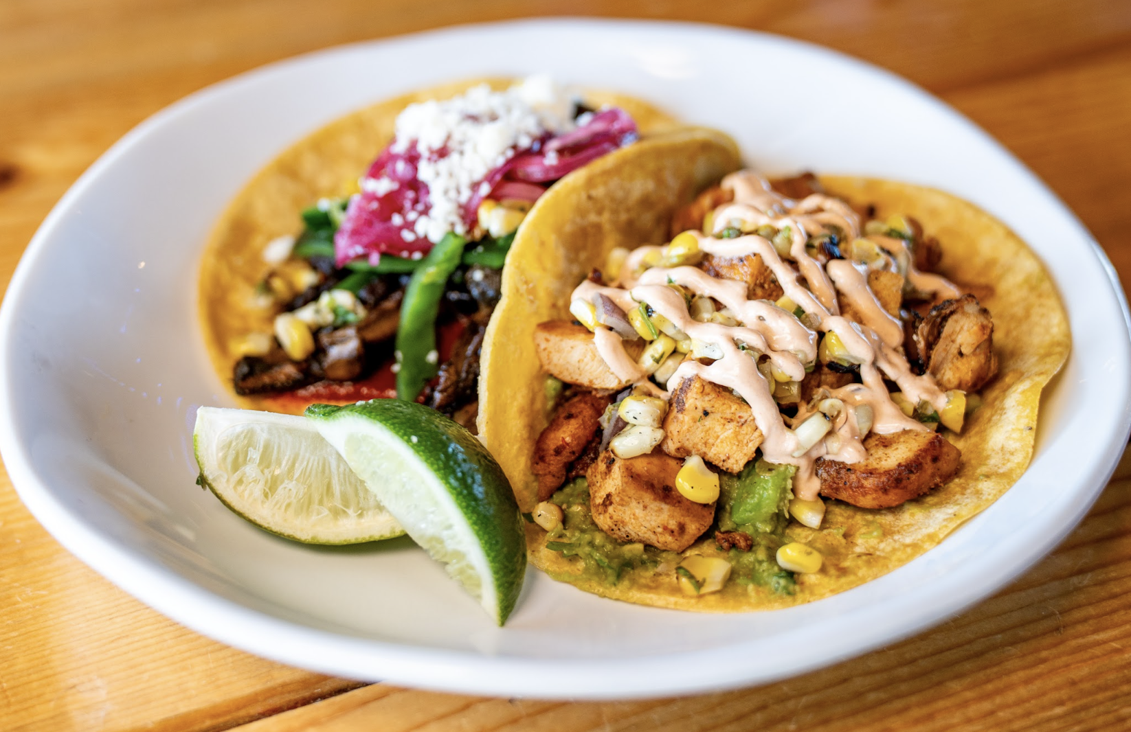 Two tacos on a white plate: one with grilled chicken, corn, and creamy sauce, the other with vegetables and cheese. A lime wedge is on the side. The plate is set on a wooden table.
