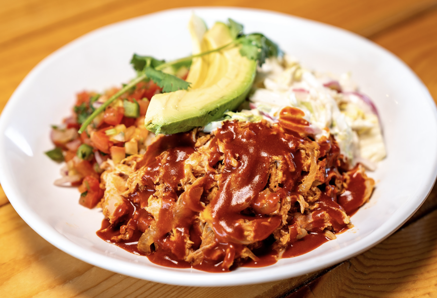 Shredded meat with rich red sauce, topped with sliced avocado and cilantro, served with pico de gallo and creamy coleslaw on a white plate.
