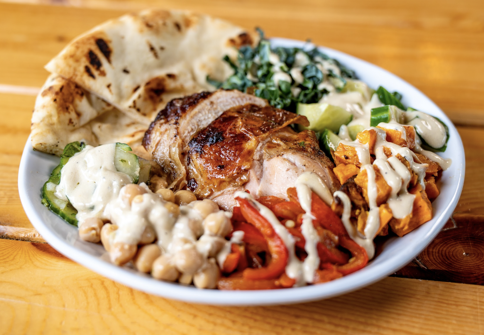 A plate with grilled chicken, pita bread, sautéed greens, roasted sweet potatoes, chickpeas, cucumbers, and red peppers, all drizzled with a creamy sauce, on a wooden table.