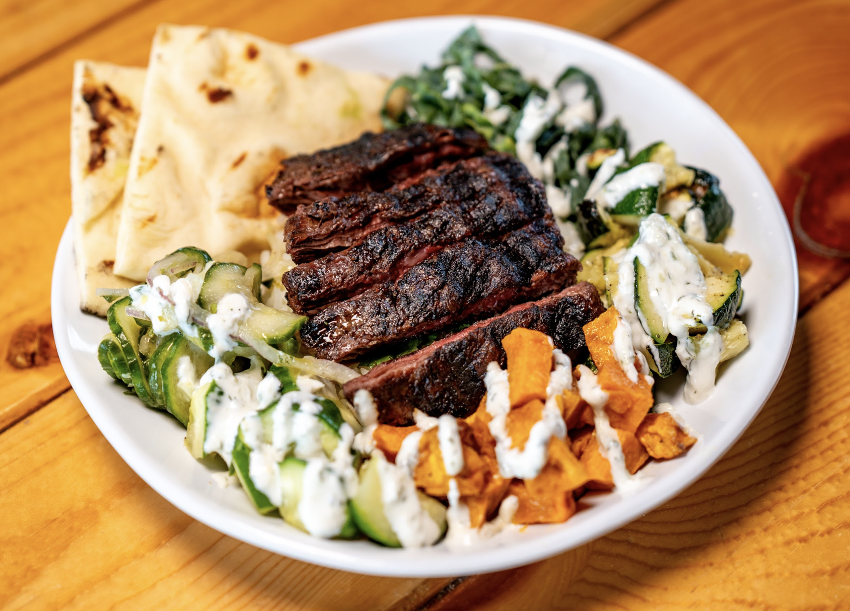 A plate with sliced grilled steak, naan bread, cucumber, sweet potatoes, greens, zucchini, and tzatziki sauce drizzled on top, served on a wooden table.