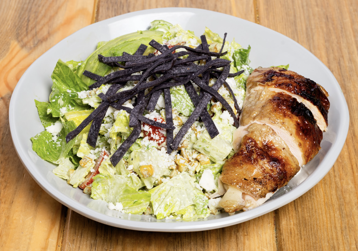 A plate with sliced roasted chicken next to a salad with lettuce, avocado, black tortilla strips, crumbled cheese, and corn, served on a wooden table.
