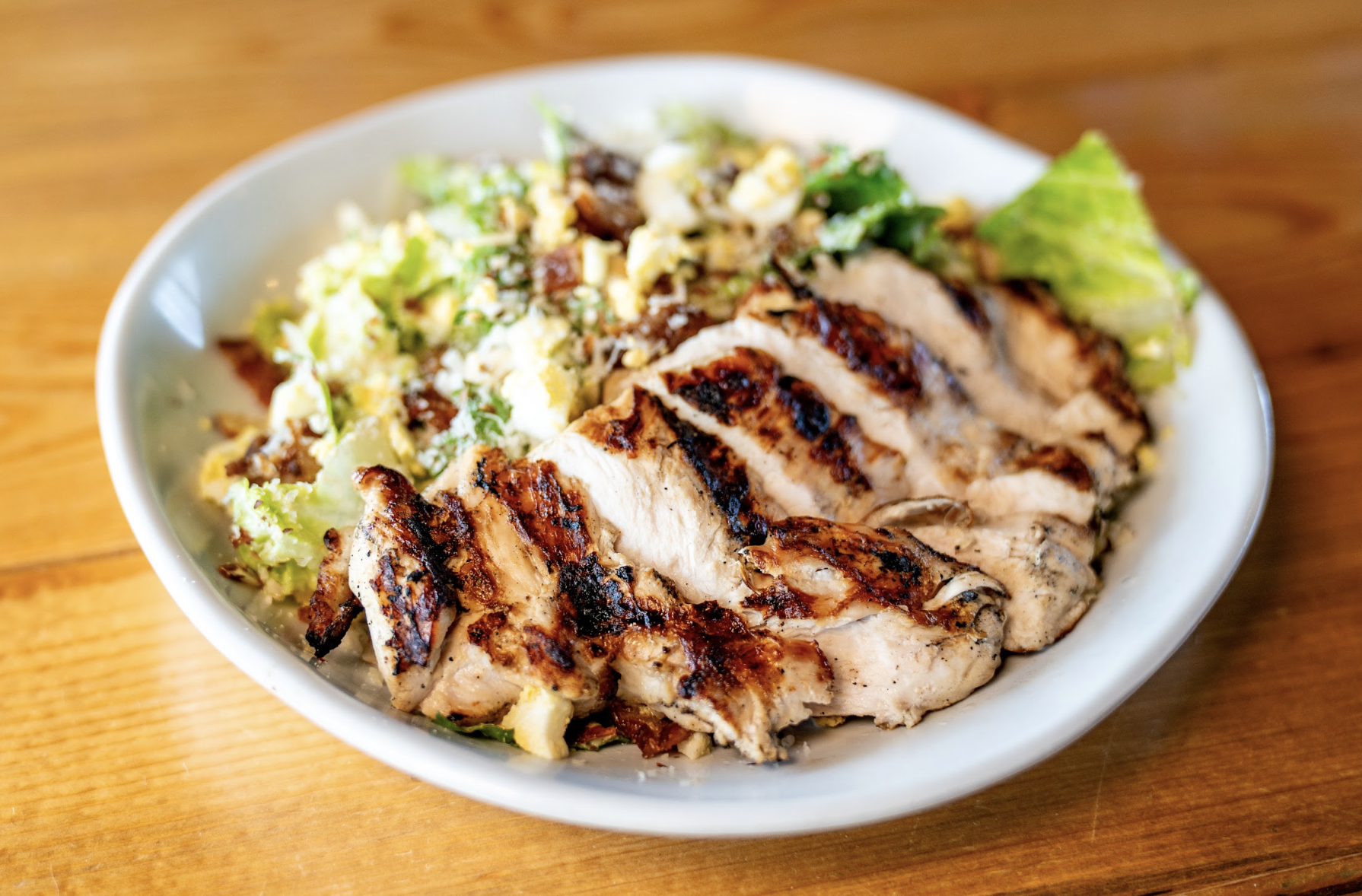 A white bowl filled with grilled chicken breast slices on top of a fresh green salad with lettuce, grated cheese, and dressing, set on a wooden table.