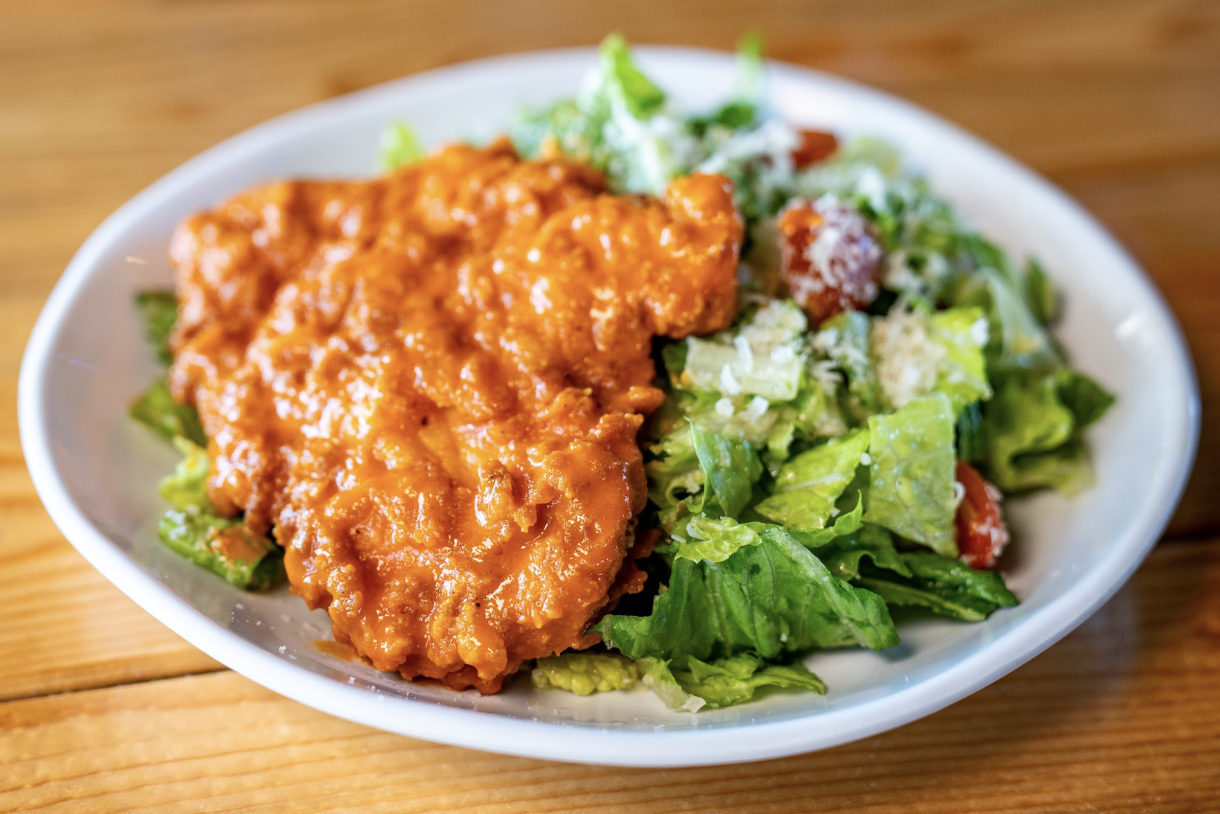 A plate of buffalo chicken on a bed of fresh green salad with tomatoes and grated cheese, served on a white dish atop a wooden table.