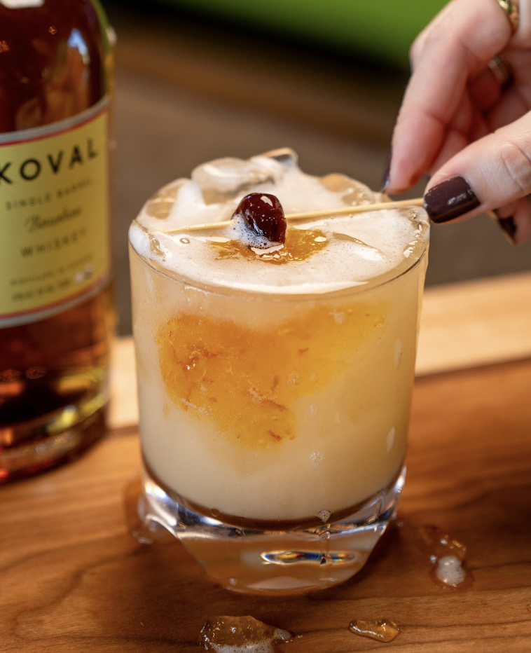 A hand garnishes a whiskey sour with a cherry. The drink sits in a glass with ice and foam on top, next to a Koval whiskey bottle on a wooden surface.