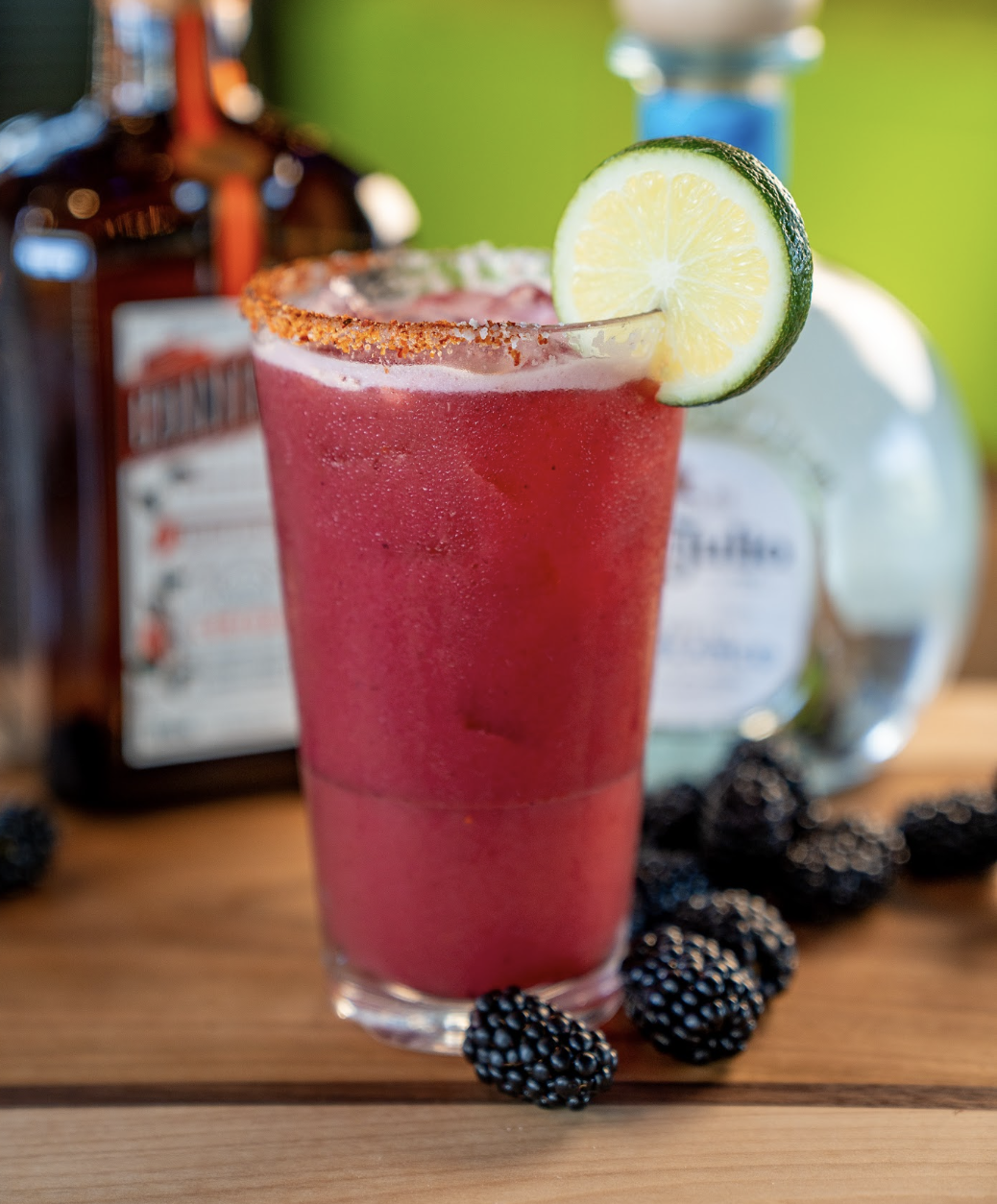 A vibrant red cocktail in a glass with a chili rim, garnished with a lime wheel. Blackberries are scattered around the base, with blurred bottles in the background.