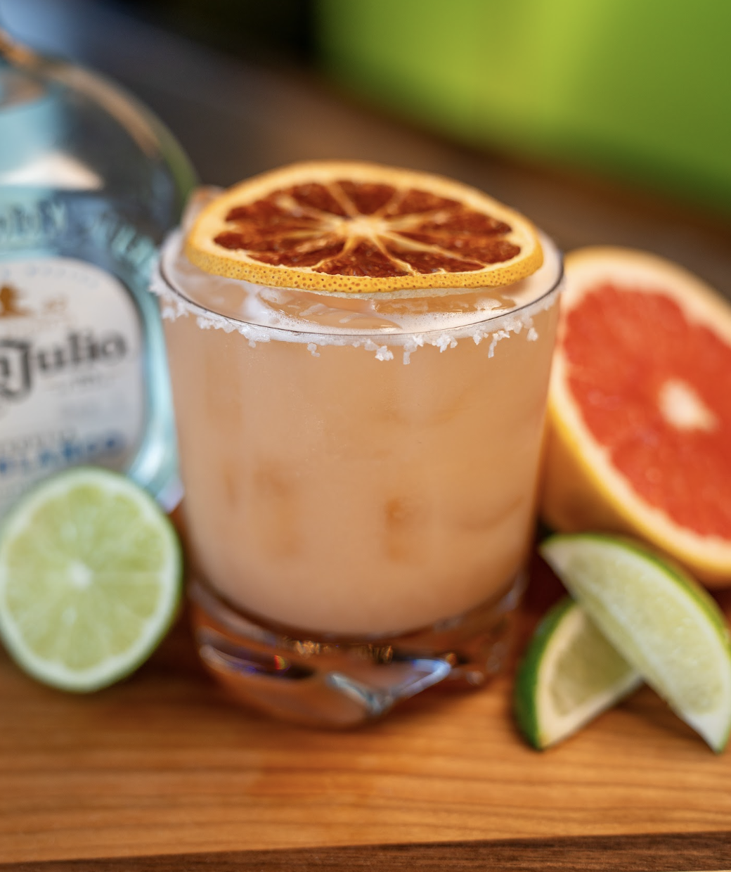 A cocktail with a salted rim and dried citrus slice garnish sits on a wooden surface, surrounded by lime wedges, half a grapefruit, and a tequila bottle.