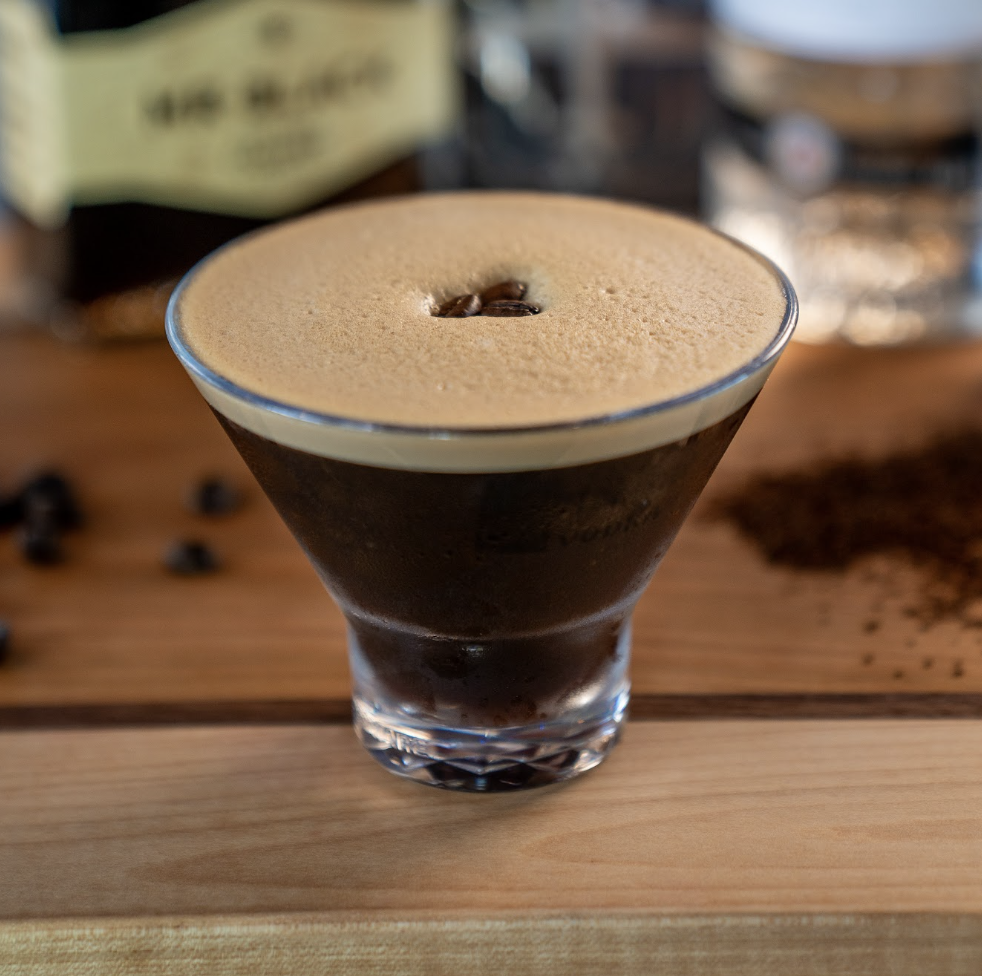 A close-up of an espresso martini in a martini glass, topped with a layer of creamy foam and a single coffee bean, set on a wooden surface with blurred bottles and coffee grounds in the background.