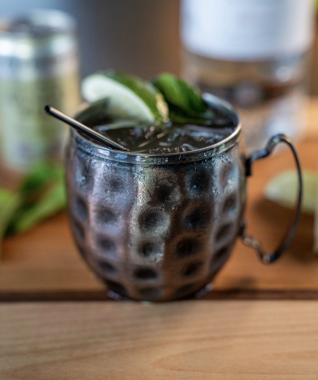 A Moscow Mule cocktail in a textured metal mug with a metal straw, garnished with lime wedges and mint, sits on a wooden surface.