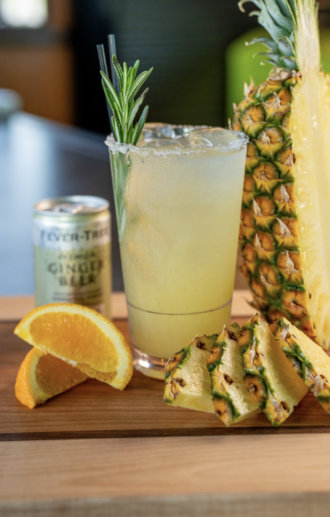 A tropical cocktail garnished with rosemary and served in a glass with a salted rim, sits beside orange slices, pineapple wedges, a half pineapple, and a can of ginger beer on a wooden board.