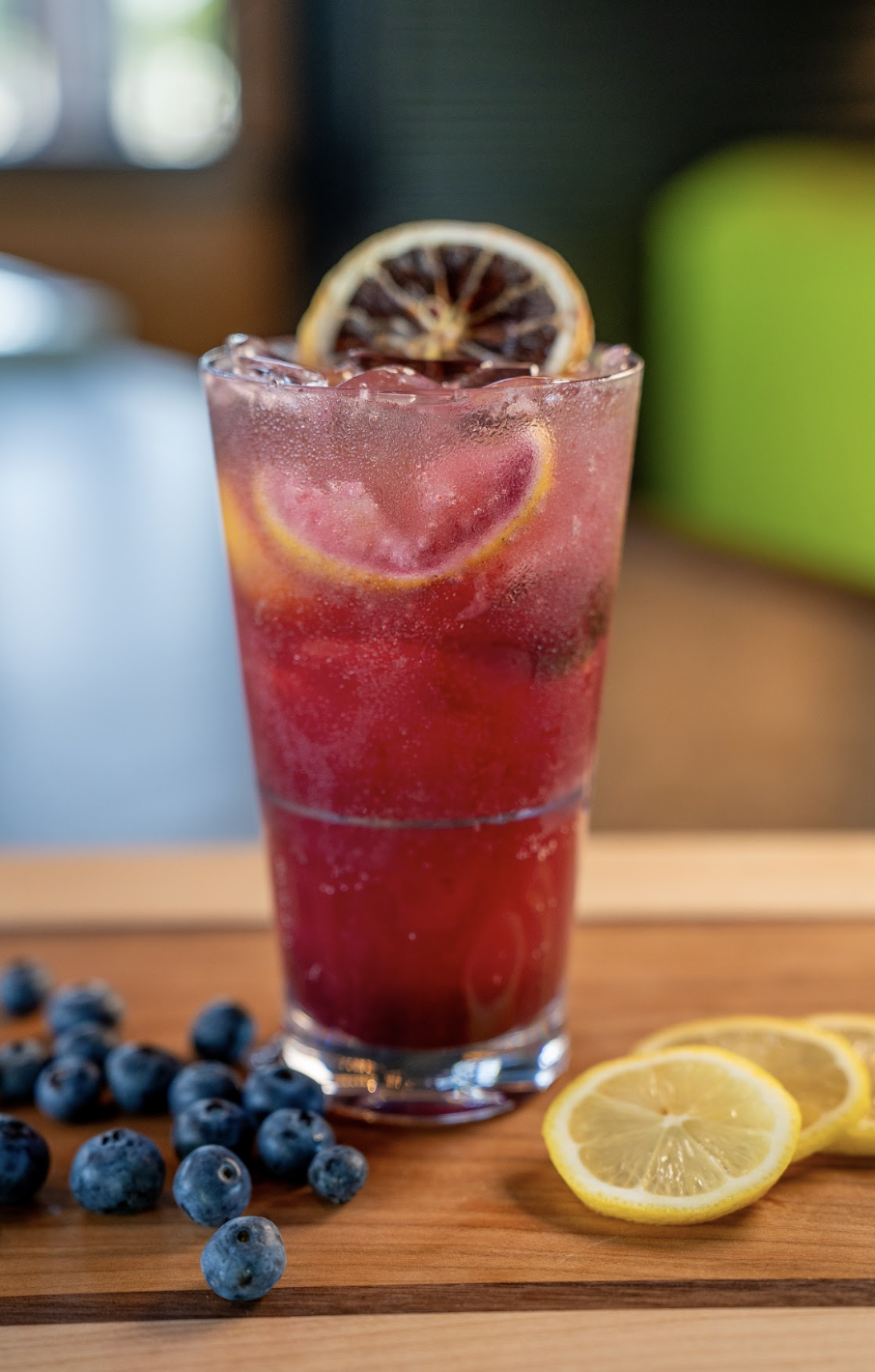 A glass of purple blueberry lemonade with ice, garnished with lemon slices and a dried lemon wheel, sits on a wooden surface beside fresh blueberries and lemon slices.