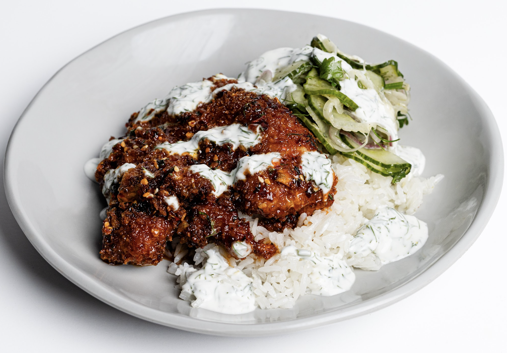 A plate of white rice topped with sauced, crispy chicken, creamy white herb sauce, and a side of cucumber salad with onions, served on a light gray plate.