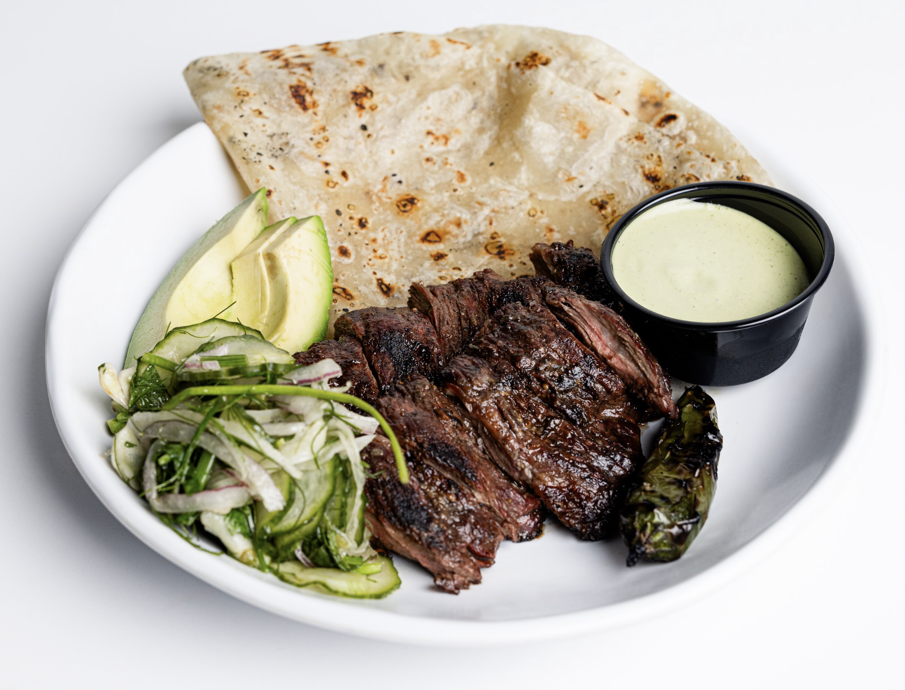 A white plate with grilled steak slices, a charred pepper, sliced avocado, mixed green salad, a folded flatbread, and a small cup of green sauce.