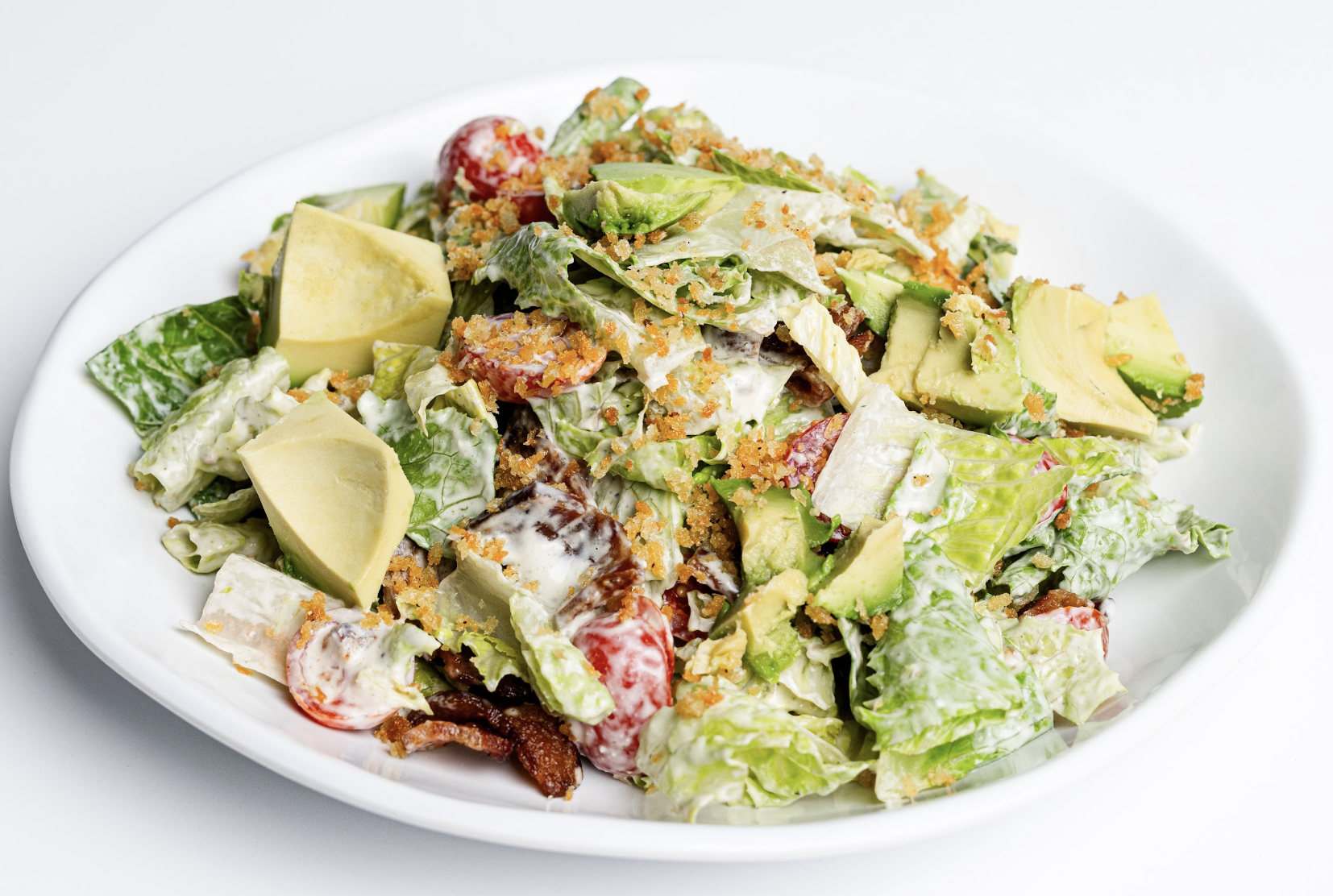 A white plate with a fresh salad containing lettuce, cherry tomatoes, avocado chunks, and crispy toppings, all mixed with a creamy dressing.