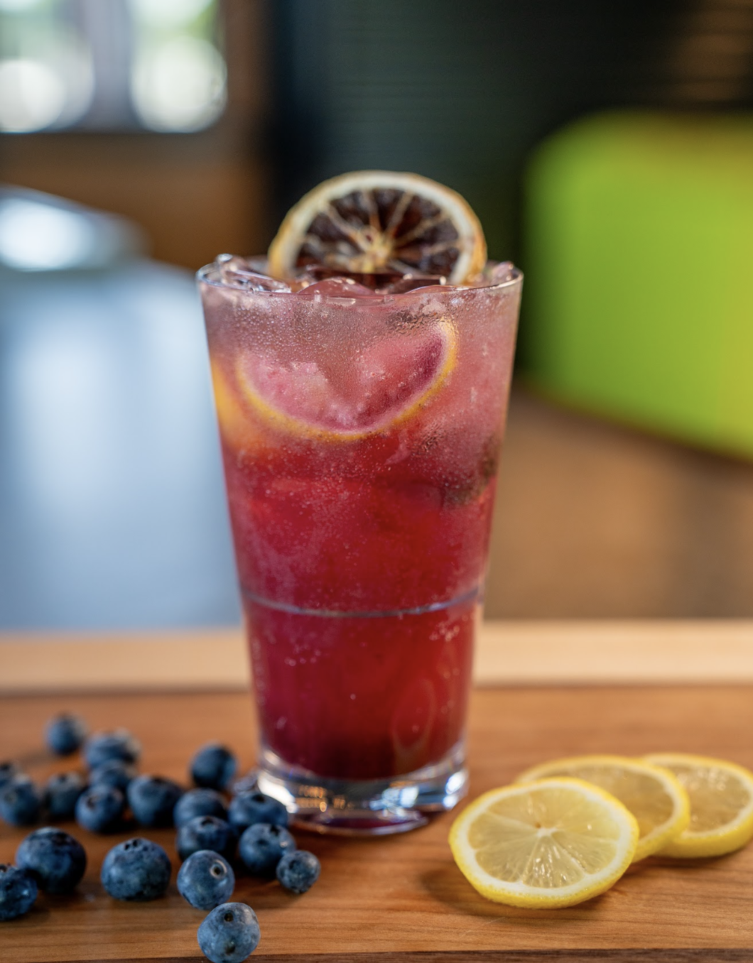 A tall glass of pink lemonade with ice, lemon slices, and a dried lemon garnish sits on a wooden surface next to fresh blueberries and lemon slices. The background is softly blurred.