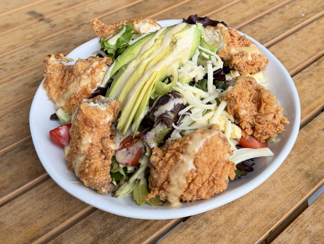 A white bowl filled with fried chicken strips, sliced avocado, shredded cheese, mixed greens, tomatoes, and dressing from our North Kansas City Menu sits on a wooden table.