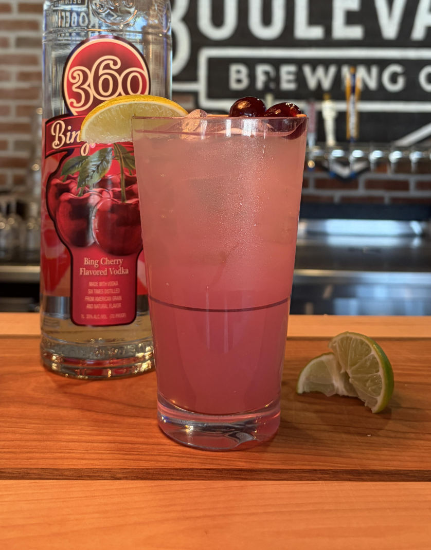 A pink cocktail garnished with lime and cherries sits on a bar beside a bottle of 360 Bing Cherry Flavored Vodka, with lime wedges on the wooden counter—just one tempting highlight from the North Kansas City Menu.