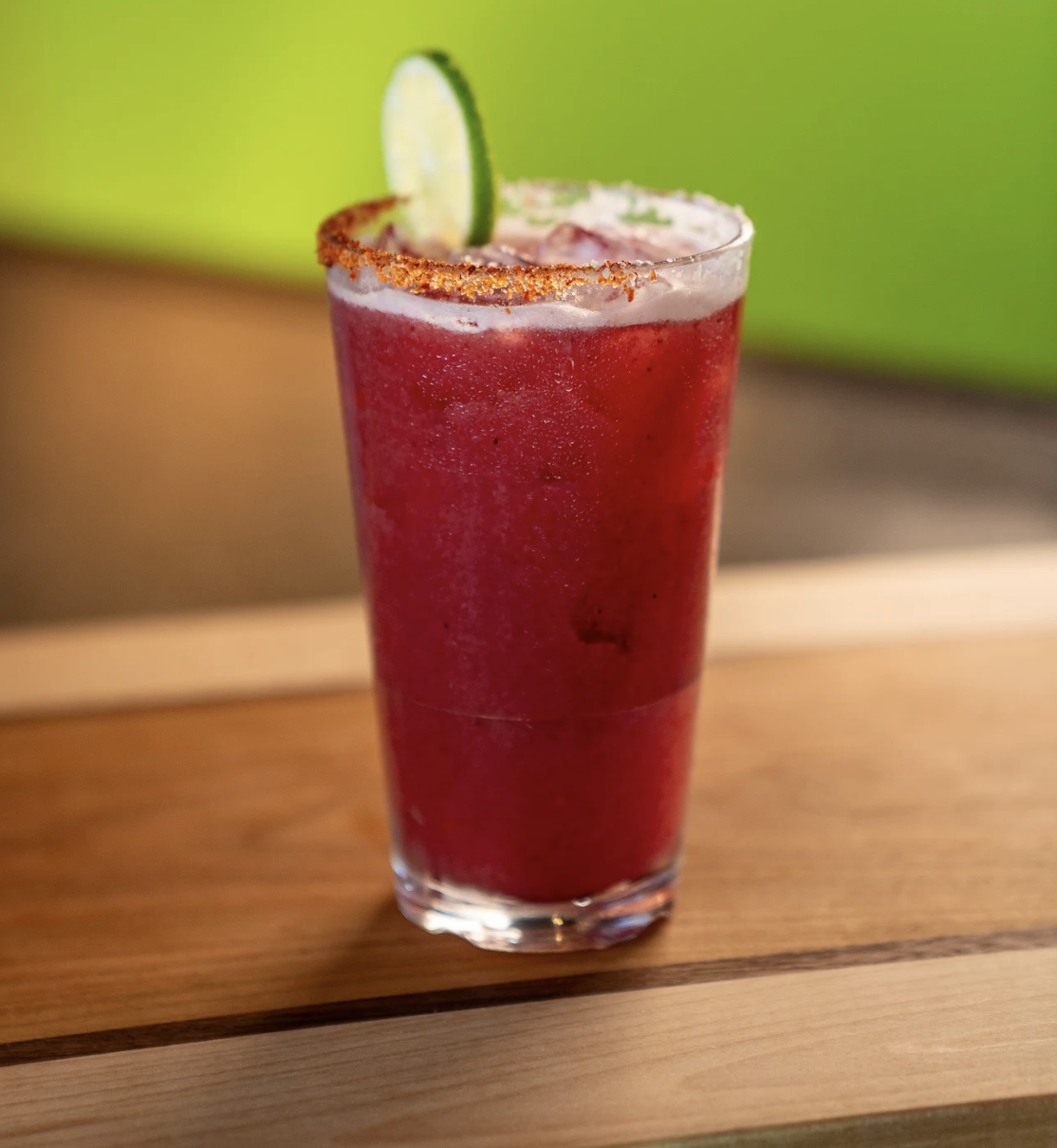 A vibrant red slushy drink from the North Kansas City Menu sits in a clear glass, topped with a lime wheel and chili powder on the rim, all resting on a wooden surface with a lush green background.
