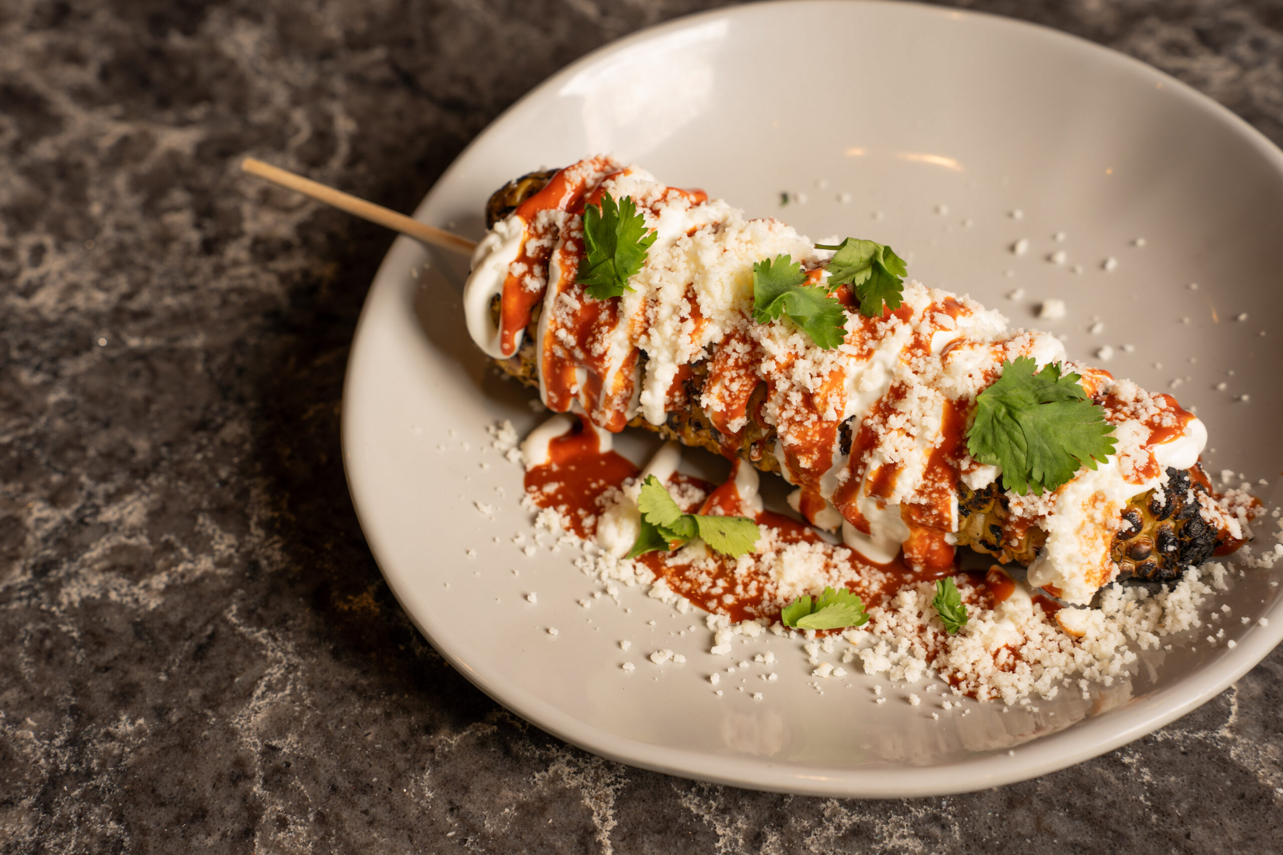 A plate with Mexican street corn on a stick, topped with creamy sauce, cheese, red chili sauce, and fresh cilantro, set on a dark stone countertop.