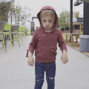 A child in a torn red hoodie and distressed jeans stands outdoors on a sidewalk, appearing to be dressed as a zombie, with pale makeup and dark circles under their eyes.