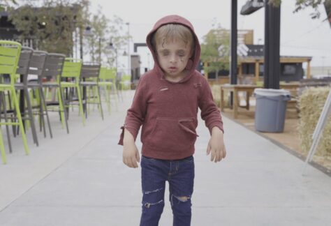 A child in a torn red hoodie and distressed jeans stands outdoors on a sidewalk, appearing to be dressed as a zombie, with pale makeup and dark circles under their eyes.