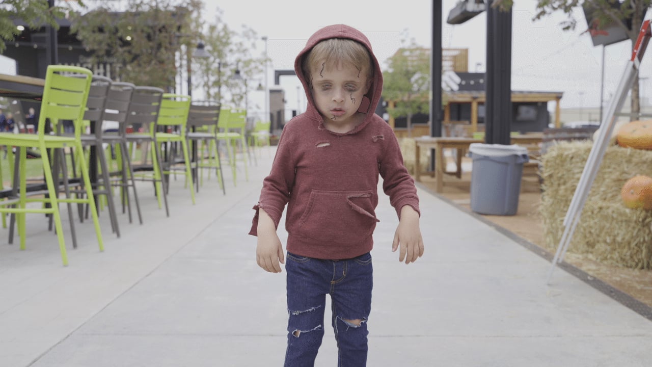 A child in a torn red hoodie and distressed jeans stands outdoors on a sidewalk, appearing to be dressed as a zombie, with pale makeup and dark circles under their eyes.