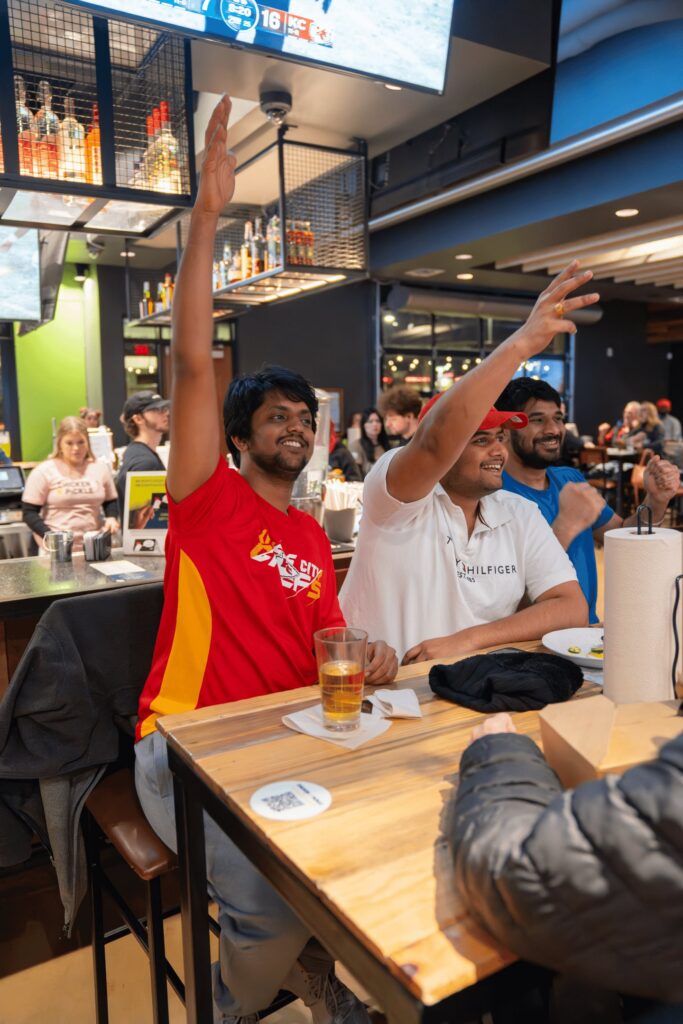 Two men sitting at a bar table inside a busy restaurant raise their hands eagerly, smiling. One wears a red shirt, the other white. Drinks and food are on the table, with people and TVs in the background.