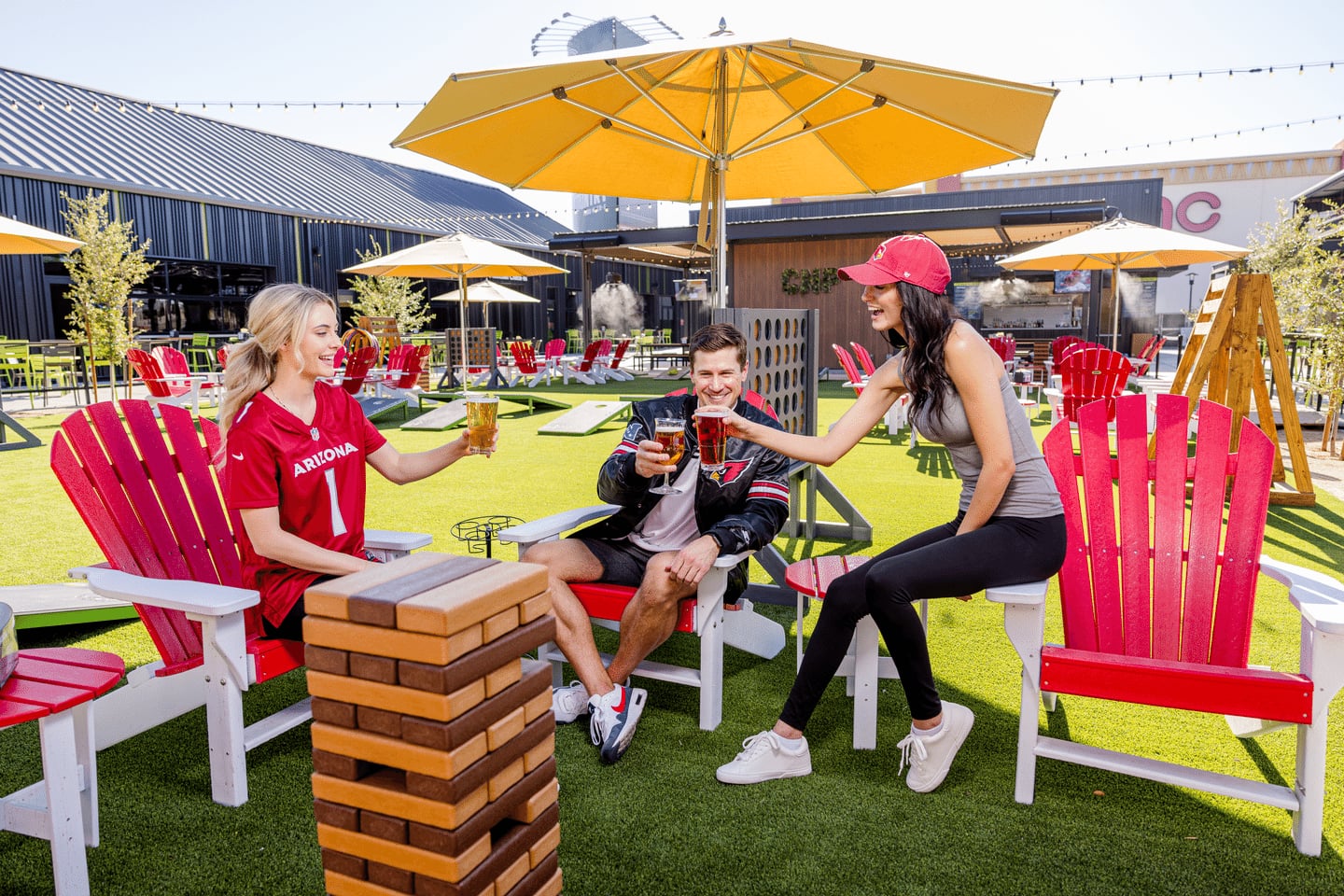 Three people sit on red Adirondack chairs outdoors, toasting drinks and smiling. There are yellow umbrellas, string lights, and a Jenga game on a grassy area with a relaxed, sunny atmosphere.