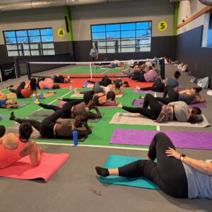 A group of people perform stretches on yoga mats in an indoor gym, with a tennis net in the background and numbered signs on the walls. Water bottles are beside the participants.