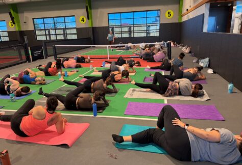 A group of people perform stretches on yoga mats in an indoor gym, with a tennis net in the background and numbered signs on the walls. Water bottles are beside the participants.