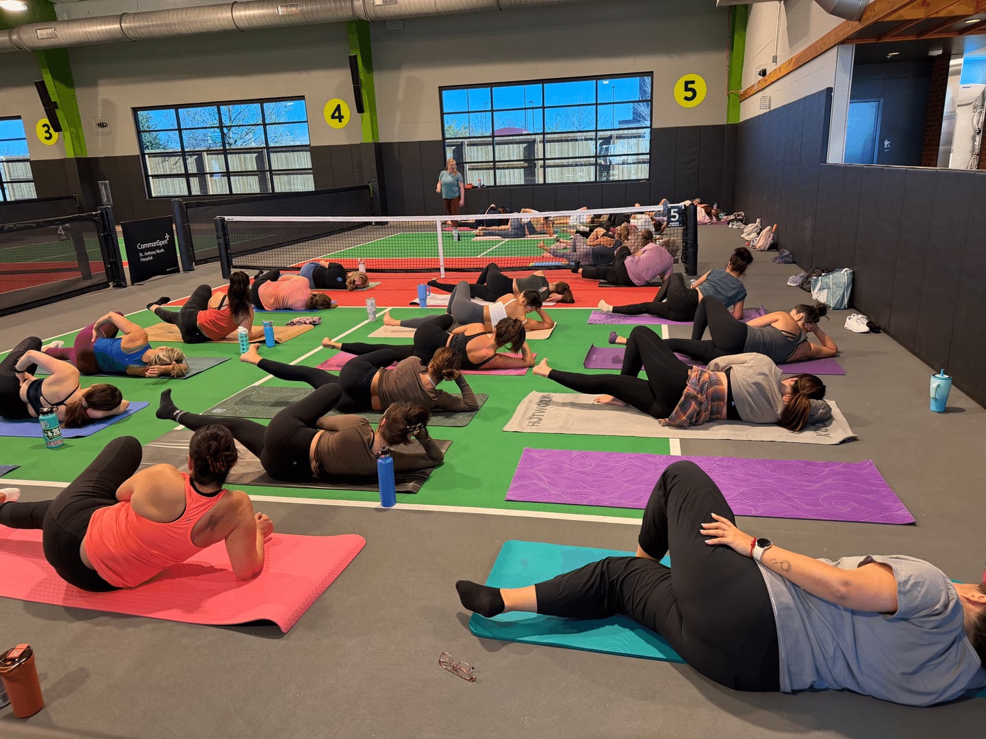 A group of people perform stretches on yoga mats in an indoor gym, with a tennis net in the background and numbered signs on the walls. Water bottles are beside the participants.