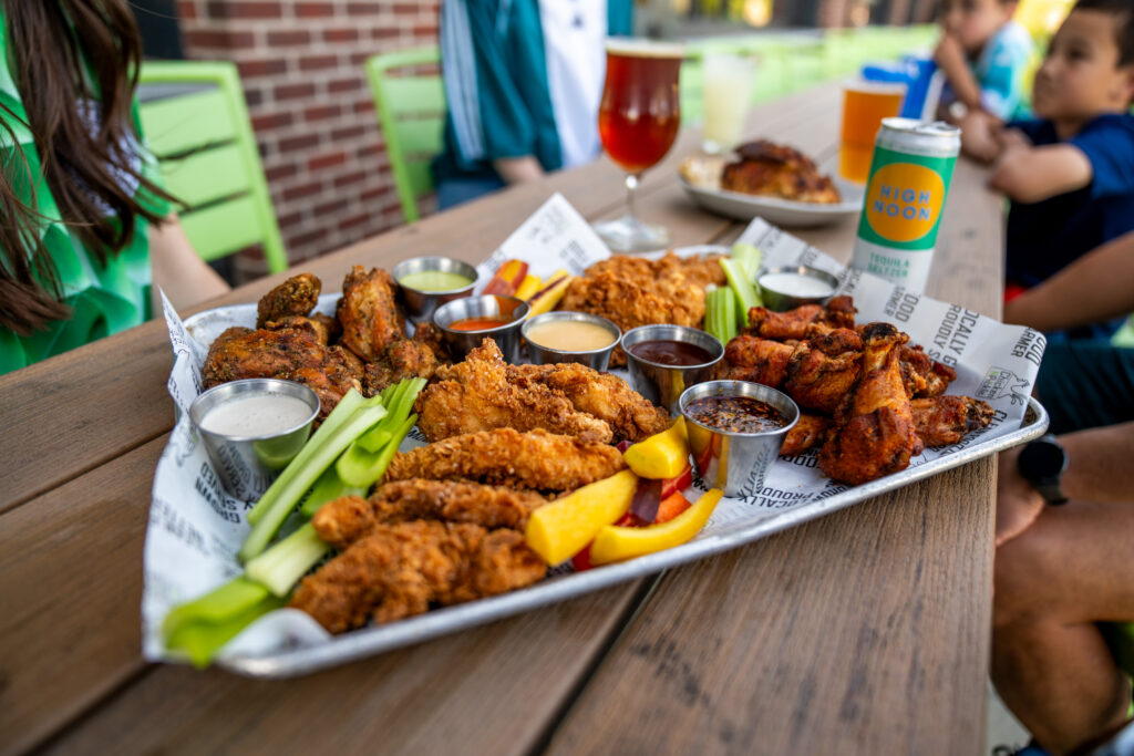 A tray of fried chicken tenders, wings, celery, and dipping sauces sits on a wooden table outside, surrounded by people. Drinks, including a can and a glass of beer, are also visible.