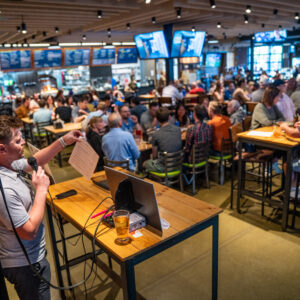 A man with a microphone hosts trivia night at a crowded bar. People sit at tables and the bar, with drinks and food, watching multiple screens under bright lights and modern décor.