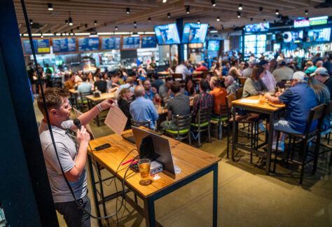 A man with a microphone hosts trivia night at a crowded bar. People sit at tables and the bar, with drinks and food, watching multiple screens under bright lights and modern décor.