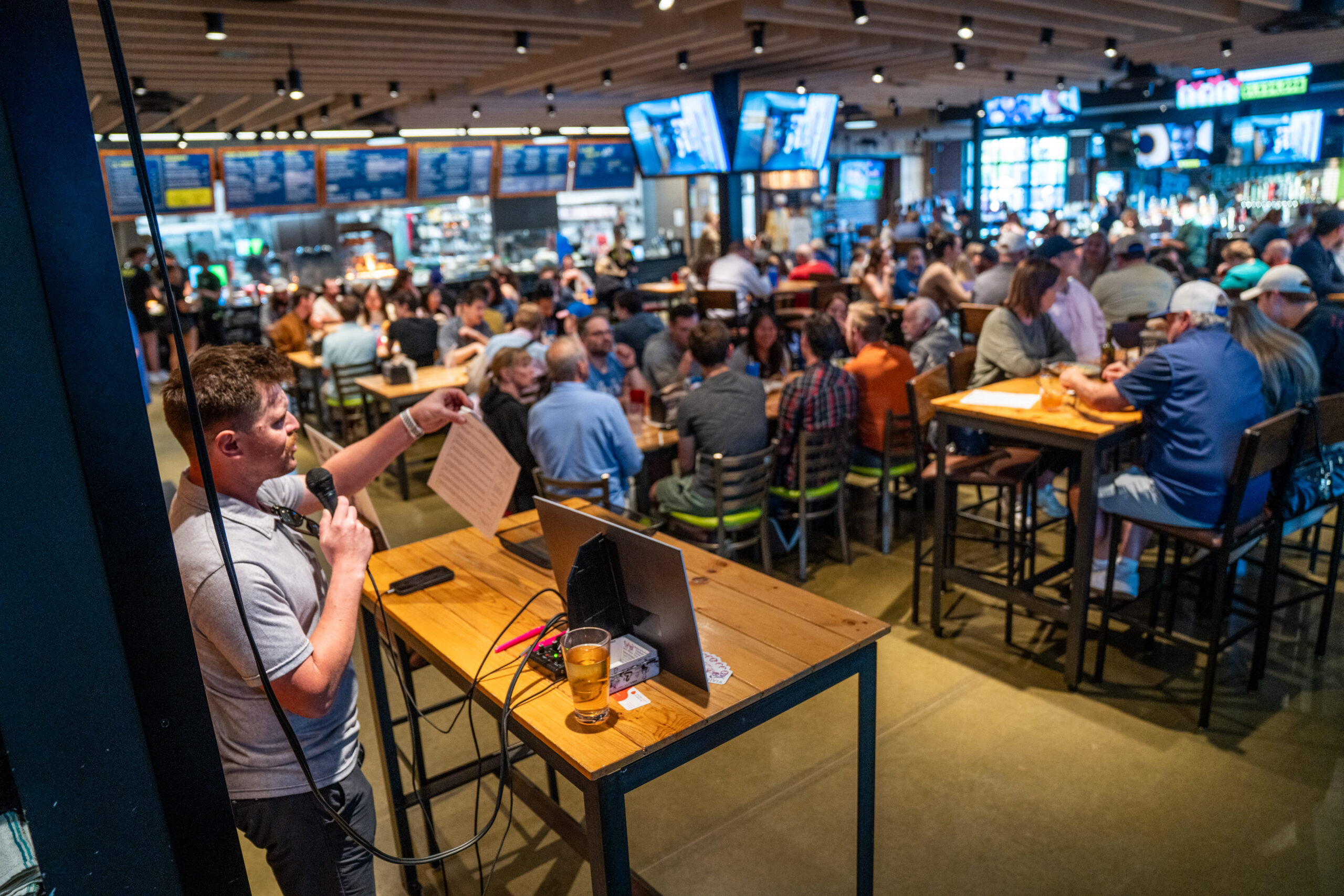 A man with a microphone hosts trivia night at a crowded bar. People sit at tables and the bar, with drinks and food, watching multiple screens under bright lights and modern décor.