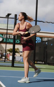 A woman in a maroon sports outfit plays pickleball on an outdoor court, holding a paddle and preparing to hit a ball. The background shows a LETT tennis sign and a partly cloudy sky.