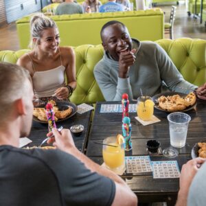 Four friends sit around a table enjoying food and drinks at a restaurant, laughing and playing bingo. A neon sign in the background reads BE AMAZING. The atmosphere is casual and lively.