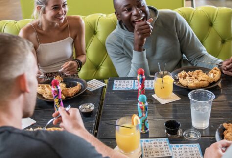 Four friends sit around a table enjoying food and drinks at a restaurant, laughing and playing bingo. A neon sign in the background reads BE AMAZING. The atmosphere is casual and lively.
