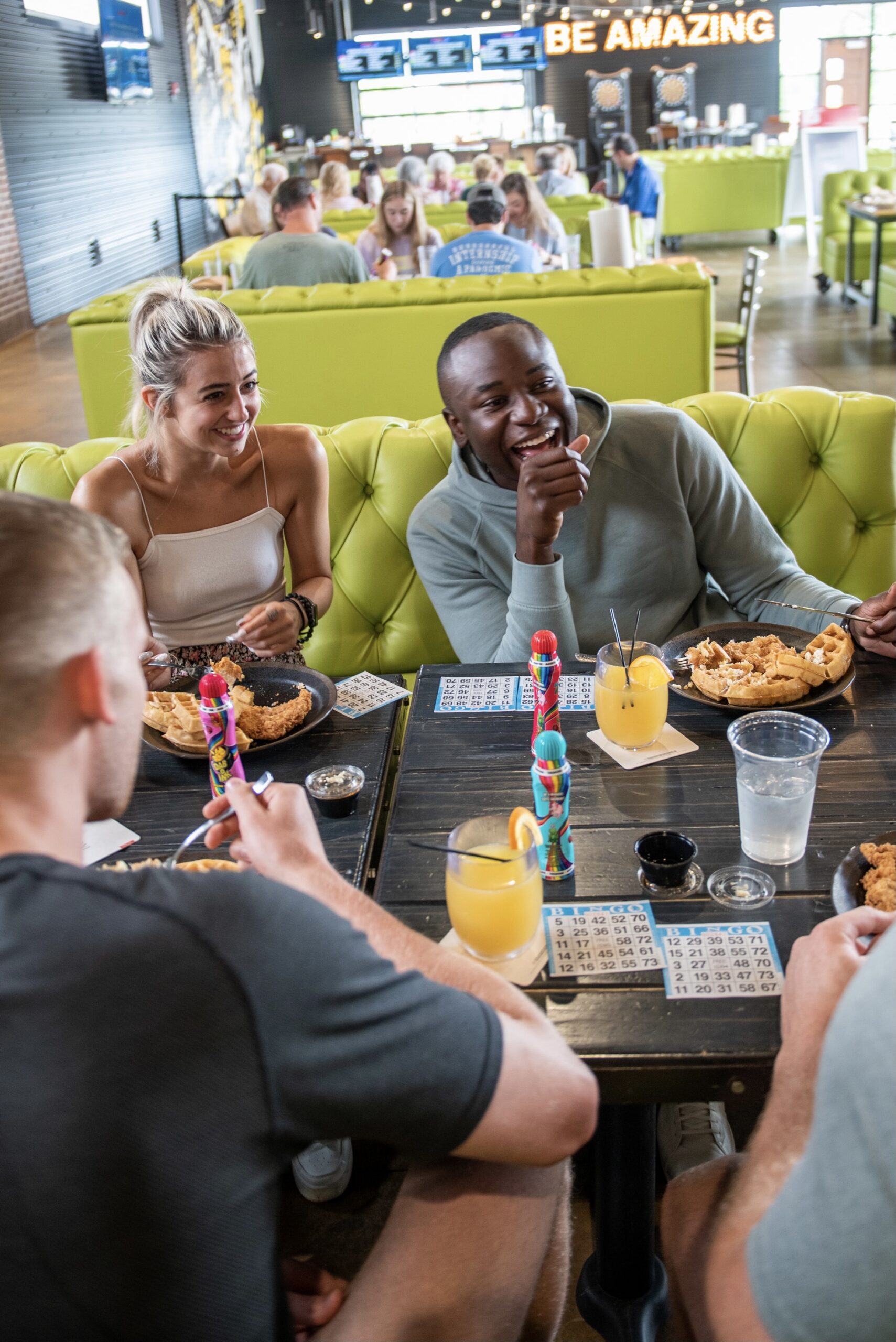 Four friends sit around a table enjoying food and drinks at a restaurant, laughing and playing bingo. A neon sign in the background reads BE AMAZING. The atmosphere is casual and lively.