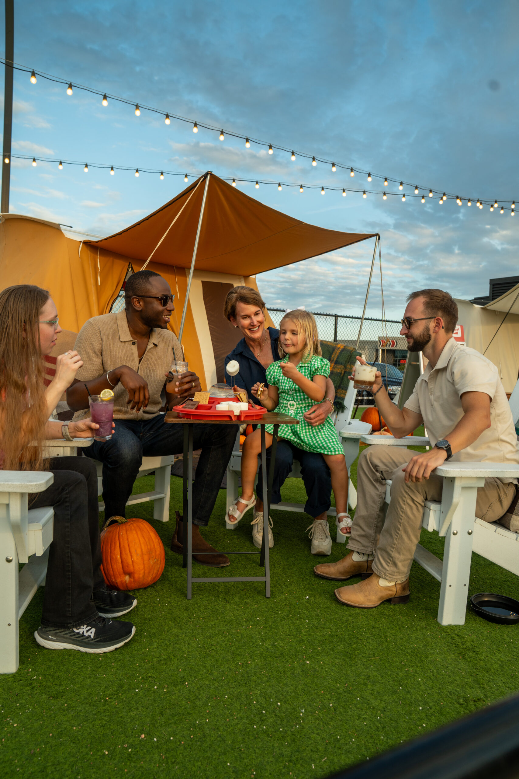 Five people, including a child, sit outdoors on white chairs around a table with snacks and drinks, under string lights near tents and pumpkins, smiling and enjoying each others company.