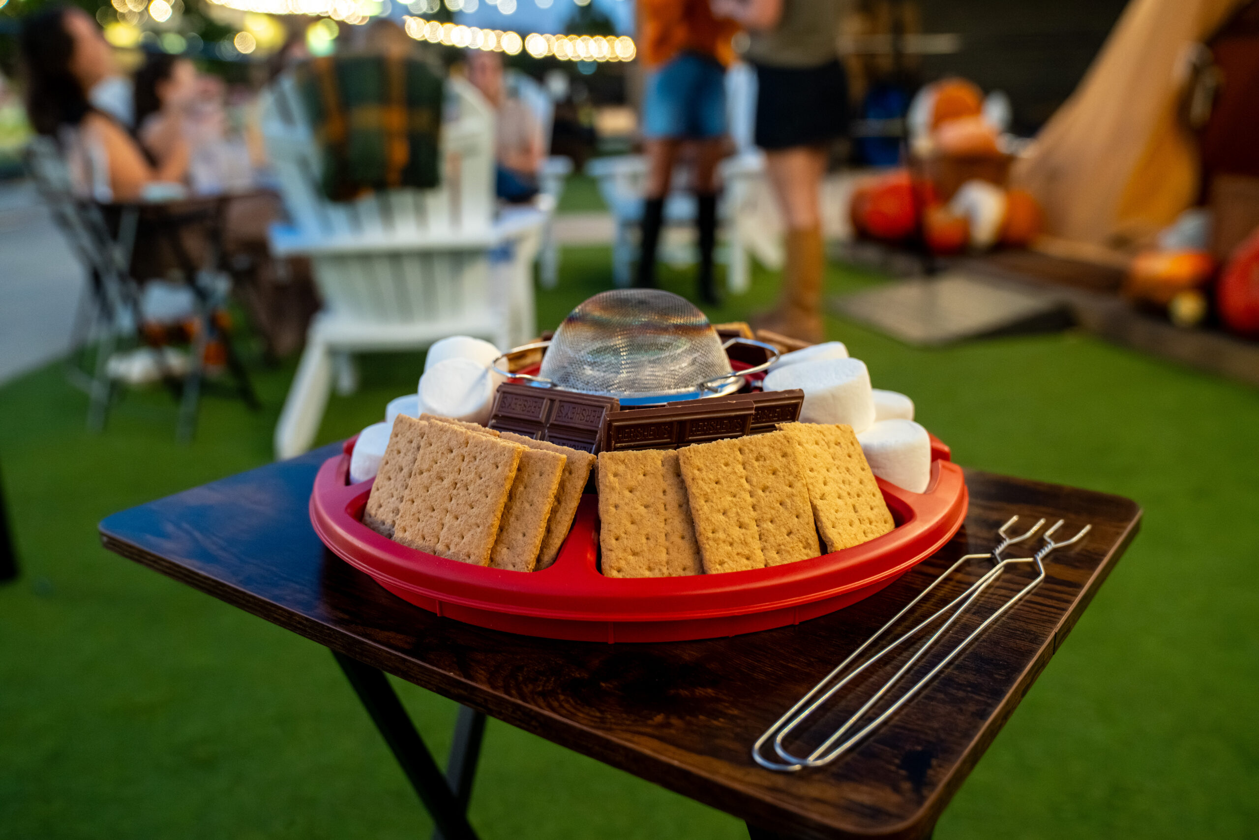 A red tray holds graham crackers, chocolate, and marshmallows for smores on a wooden table. Metal skewers are beside the tray. People and outdoor seating are blurred in the background.