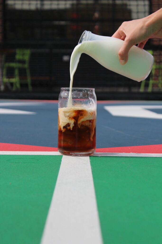 A hand pours milk from a glass bottle into a glass of iced coffee on a colorful outdoor court with green, white, blue, and red sections.
