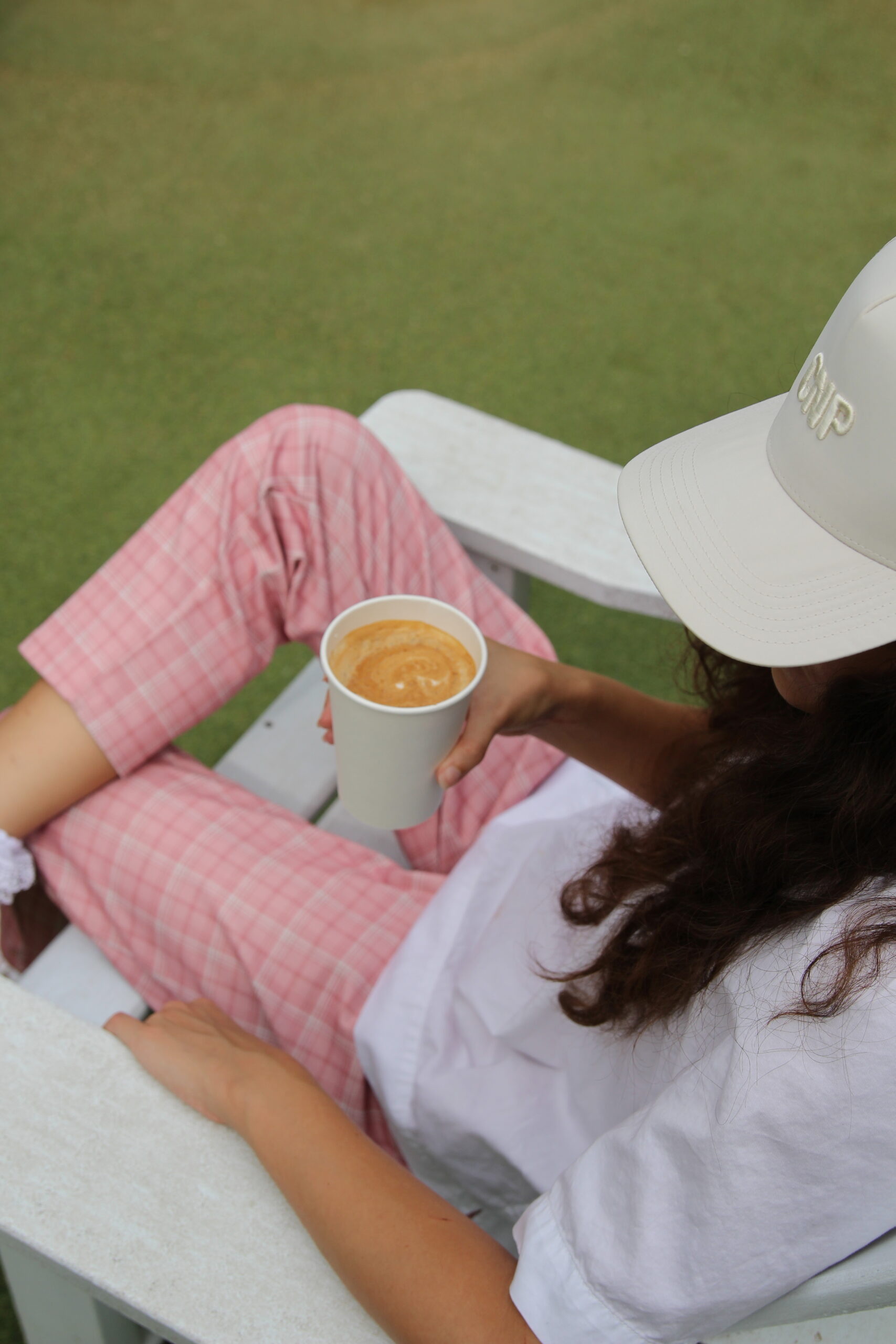 A person in pink plaid pants, a white shirt, and a white cap sits on a white chair outdoors, holding a cup of coffee.