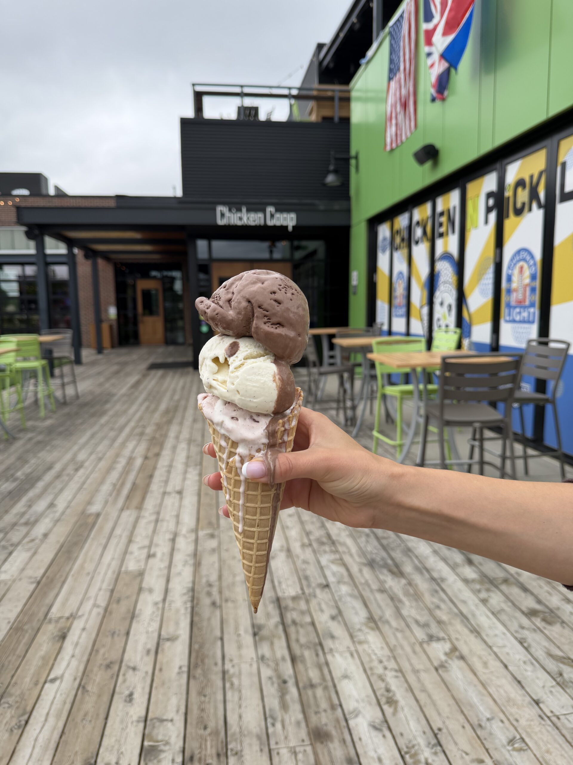 A hand holds a waffle cone with two scoops of ice cream, one chocolate and one vanilla, in front of an outdoor restaurant with colorful signs and flags.