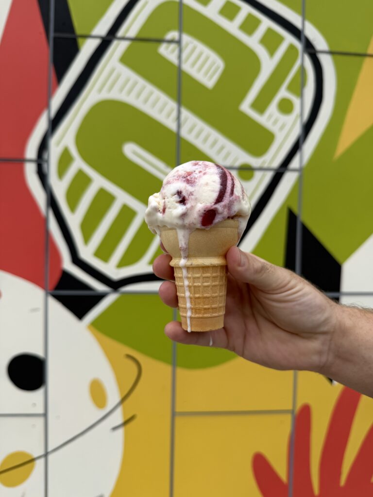 A hand holds an ice cream cone with white and red swirled ice cream in front of a colorful, abstract mural with geometric shapes and bold lines.