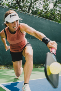 A person lunges forward on an outdoor court, focused on hitting a yellow pickleball with a paddle. They wear a visor, sleeveless top, shorts, and sneakers. Trees and a green fence are in the background.
